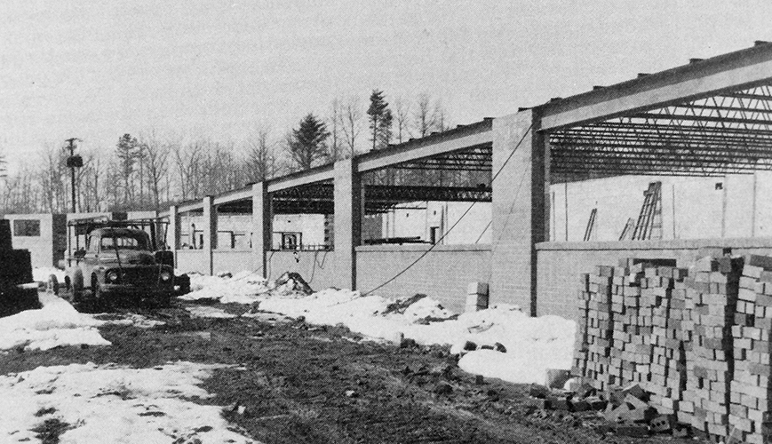 Black and white photograph of Mantua Elementary School during construction. The brick and cinderblock walls are in place, but there is no roof yet. A 1950s era pickup truck is in the distance. There is snow on the ground and a stack of bricks is in the foreground of the picture. 